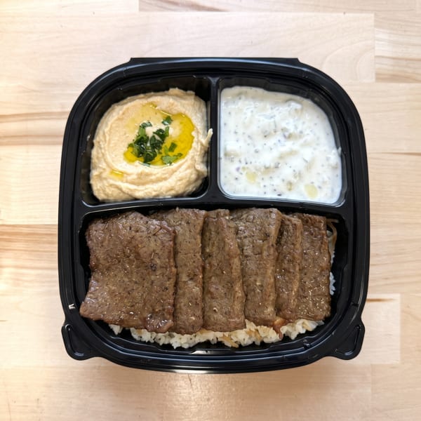 Takeout container with sliced meat over rice, hummus, and a white yogurt sauce.