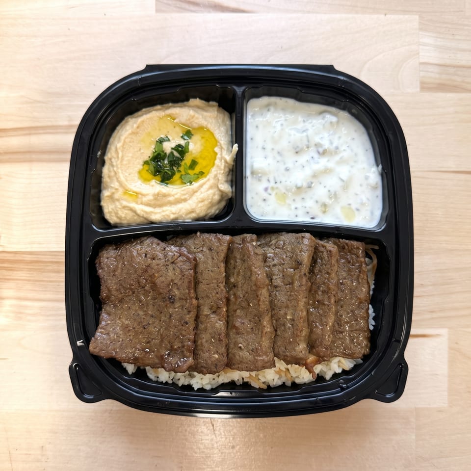Takeout container with sliced meat over rice, hummus, and a white yogurt sauce.