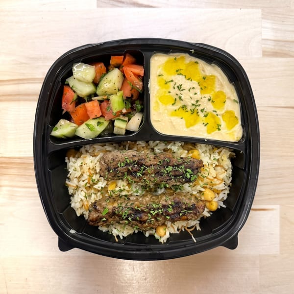 Grilled meat skewers, rice, hummus, and cucumber tomato salad in a black takeout container.