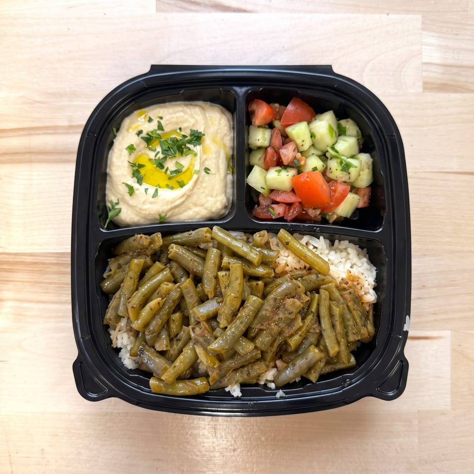 Black to-go container with hummus, cucumber tomato salad, and green beans over rice.